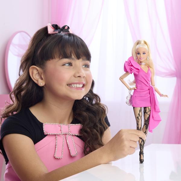 Papusa Barbie Deluxe Style impresioneaza printr-o tinuta eleganta si moderna potrivita pentru momente speciale si joaca inspirata de lumea modei Barbie poarta o rochie roz intens cu croiala asimetrica si detalii drapate completata de dresuri cu model si accesorii stralucitoare Parul blond lung coafat intr-o coada inalta completeaza look-ul sofisticat iar cerceii colierul bratara si poseta argintie adauga un plus de stil Papusa este articulata si poate fi pozitionata in diferite 