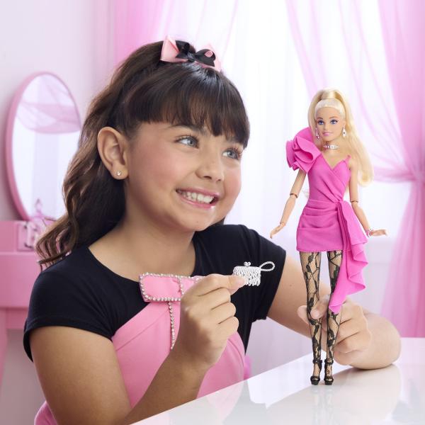 Papusa Barbie Deluxe Style impresioneaza printr-o tinuta eleganta si moderna potrivita pentru momente speciale si joaca inspirata de lumea modei Barbie poarta o rochie roz intens cu croiala asimetrica si detalii drapate completata de dresuri cu model si accesorii stralucitoare Parul blond lung coafat intr-o coada inalta completeaza look-ul sofisticat iar cerceii colierul bratara si poseta argintie adauga un plus de stil Papusa este articulata si poate fi pozitionata in diferite 